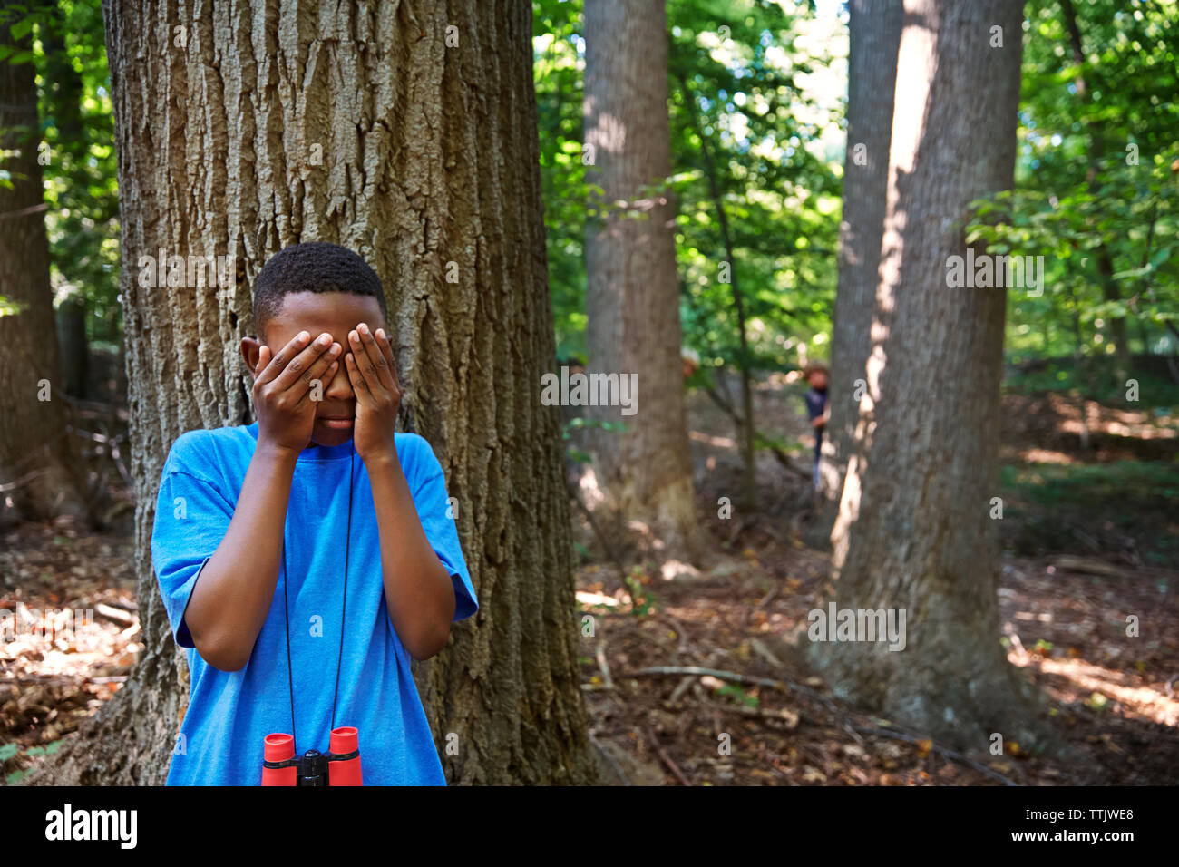 Verspielter Junge, die Augen beim Stehen gegen Baum im Wald Stockfoto