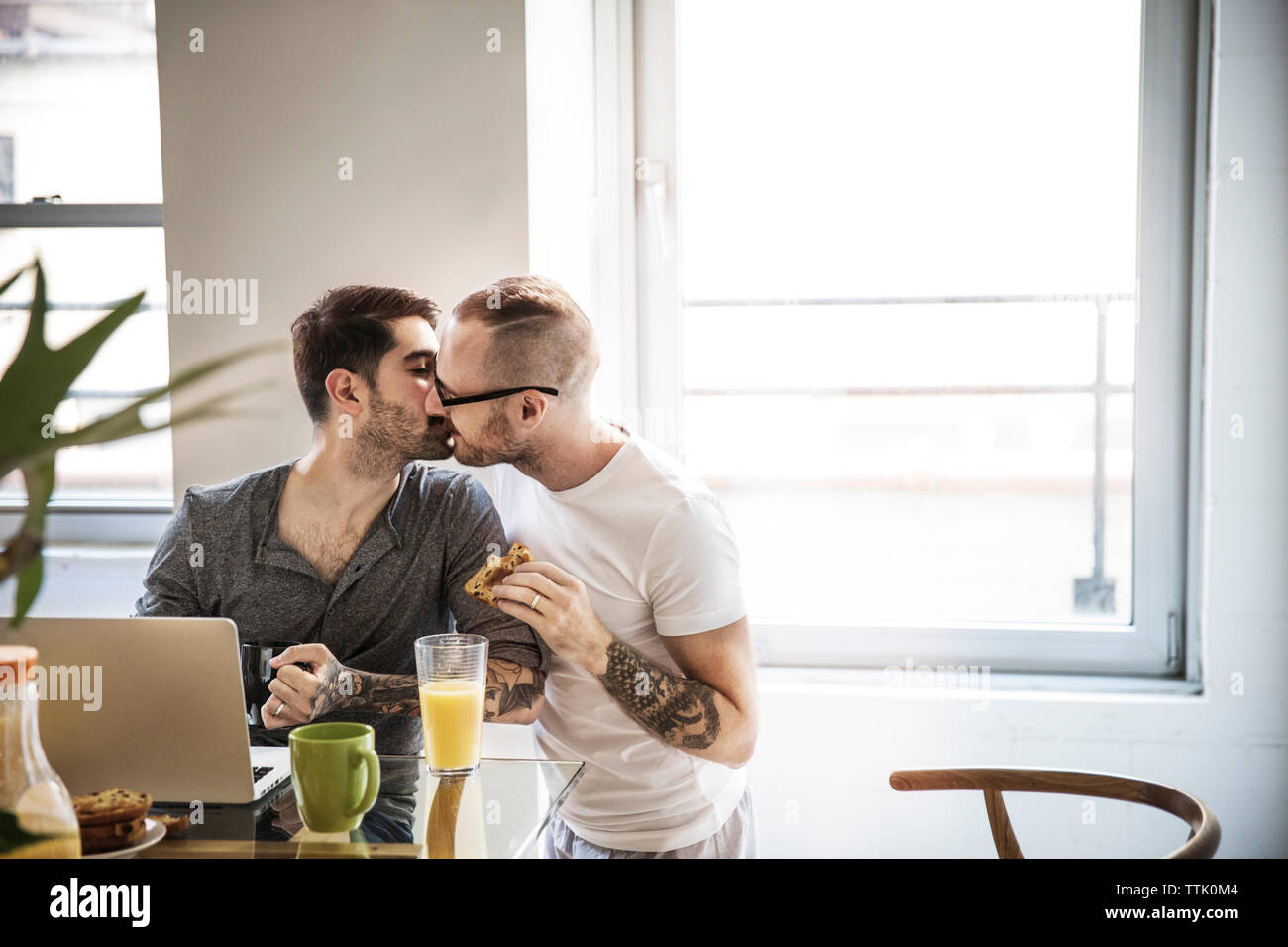 Homosexuelle Paare küssen beim Frühstück am Tisch gegen Fenster Stockfoto