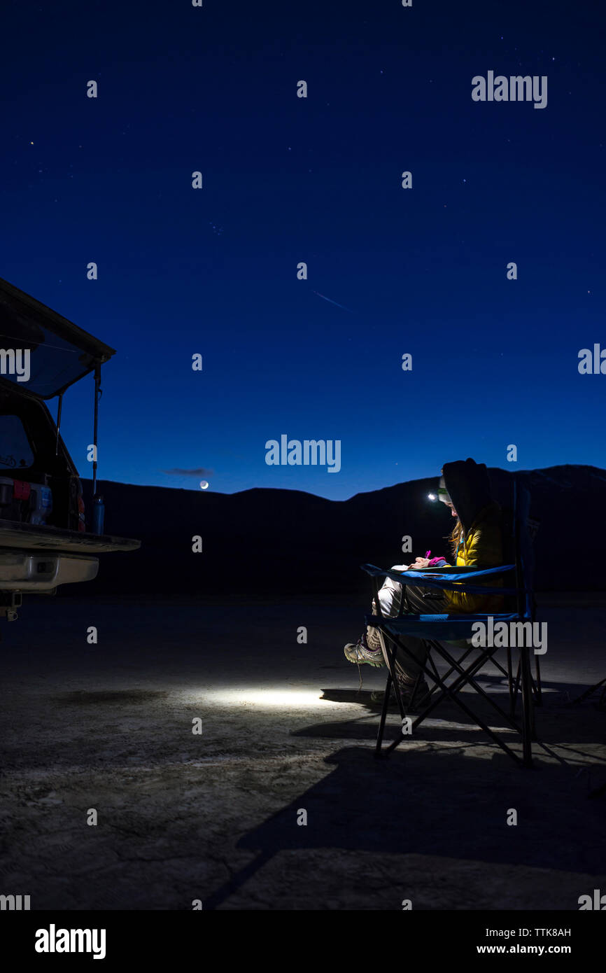 Weibliche Wanderer mit Scheinwerfer bei Nacht sitzend auf Camping Stuhl Stockfoto
