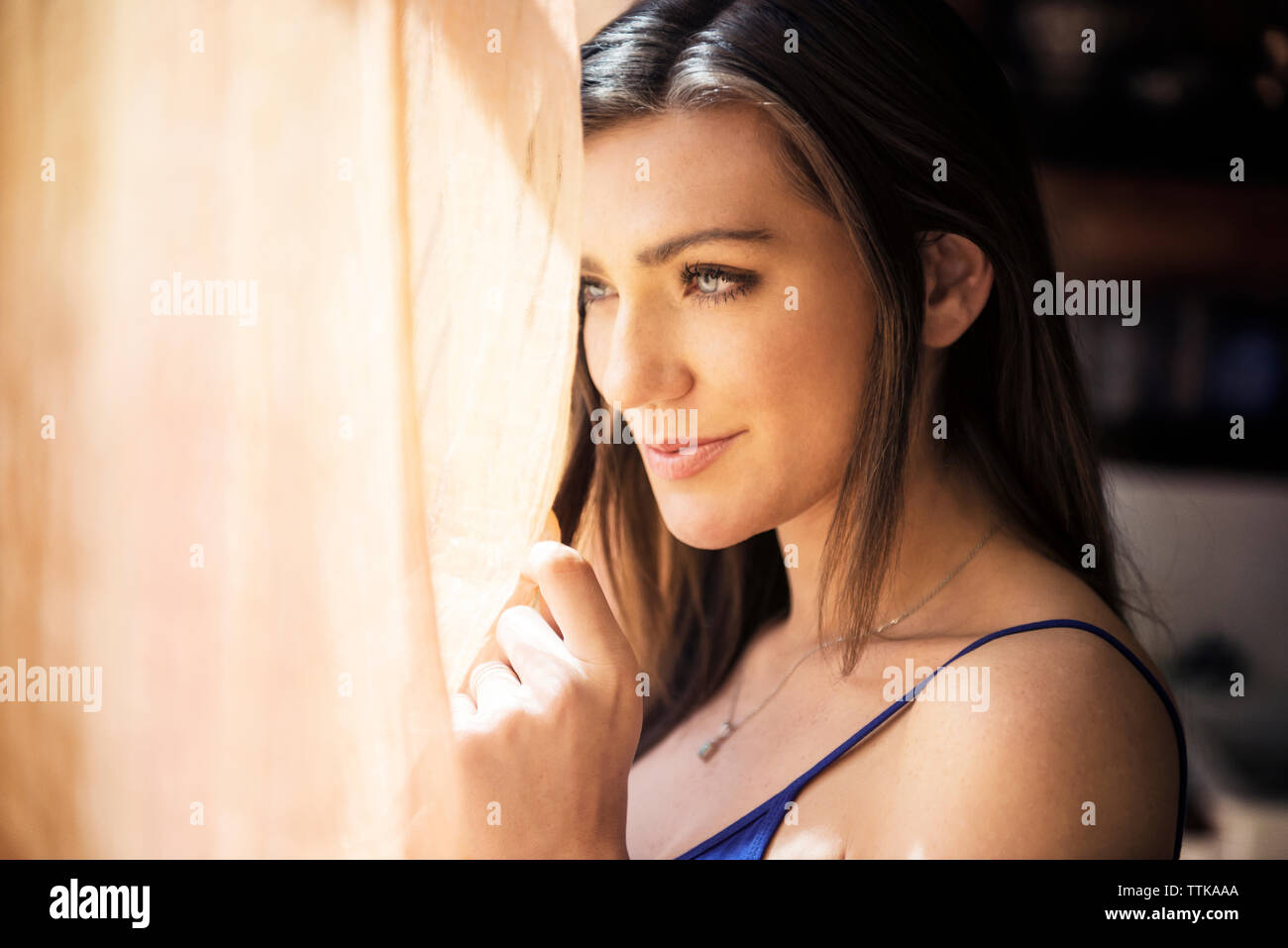 Nachdenklich lächelnde Frau stehend, durch Vorhang zu Hause Stockfoto