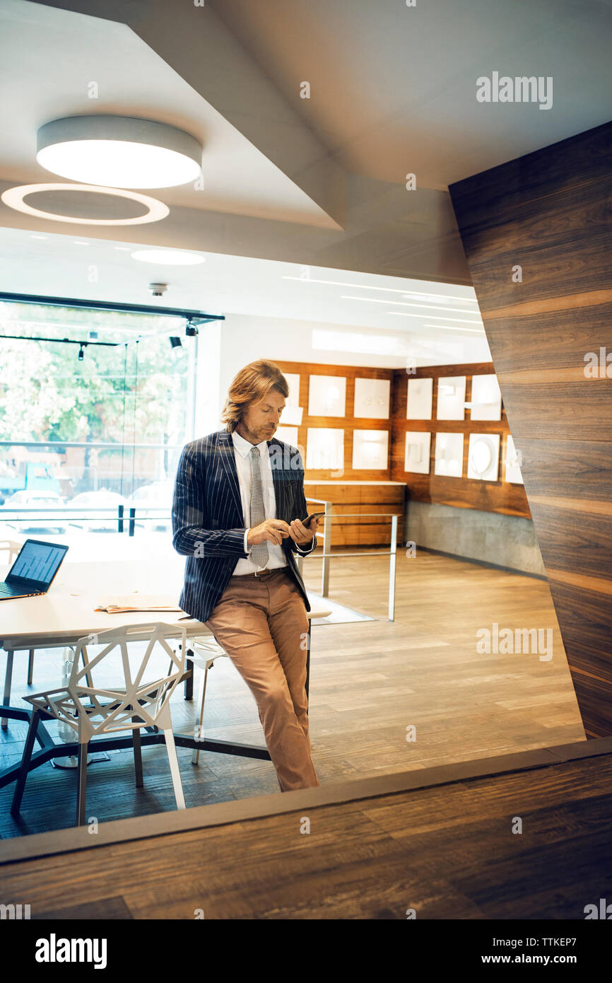 Geschäftsmann lehnte sich auf den Schreibtisch, während er sein Smartphone im Büro benutzte Stockfoto