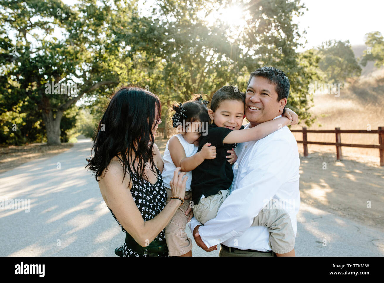 Glücklich, multi-ethnischen Eltern tragen süße Kinder, während auf der Straße gegen Bäume im Park Stockfoto