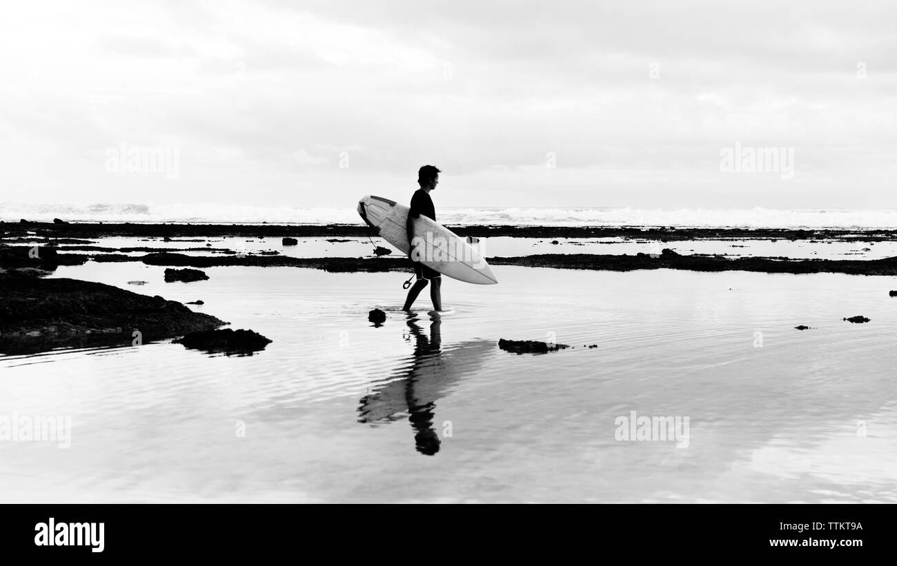 Seitenansicht der touristischen Holding Surfbrett beim Gehen auf Ufer am Strand gegen Sky Stockfoto