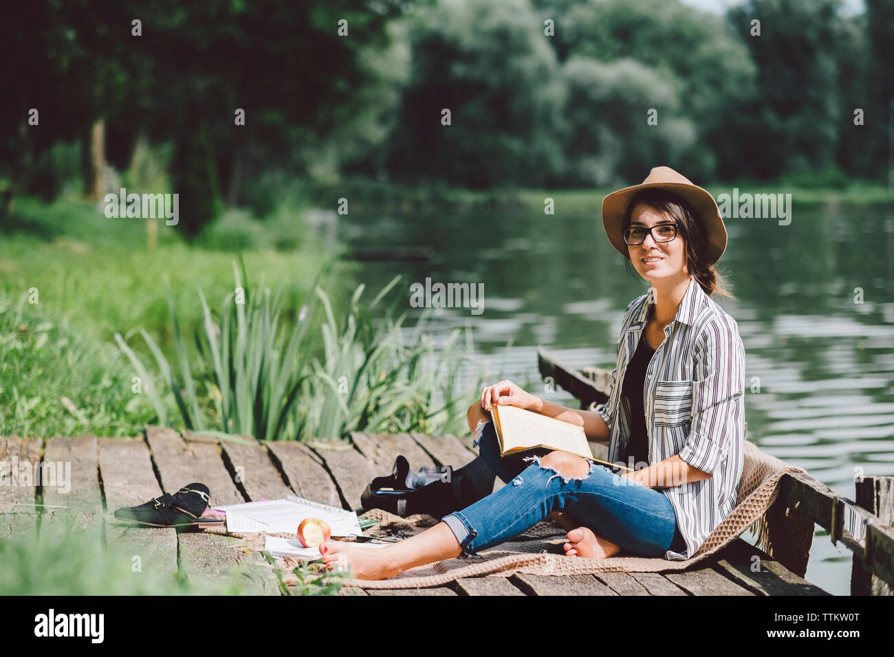Portrait von Frau mit Buch sitzen auf Pier gegen See im Wald Stockfoto