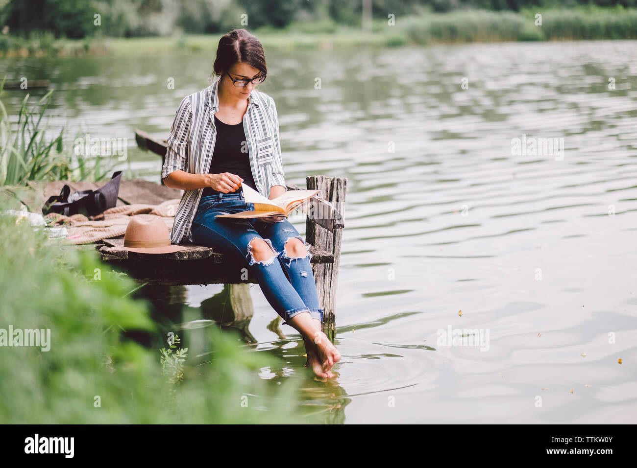 Frau mit Buch beim Sitzen auf Pier über den See im Wald Stockfoto