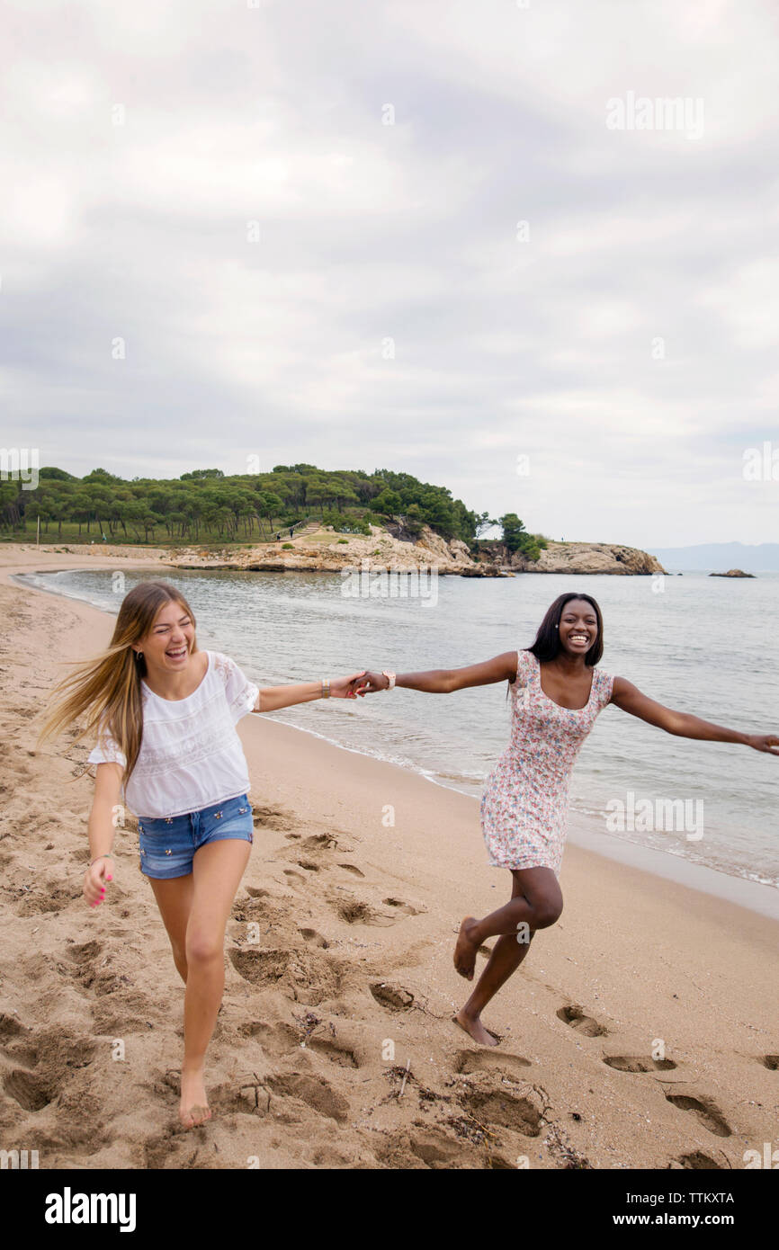 Freundliche weibliche Freunde halten sich an den Händen und Laufen am Strand gegen Sky Stockfoto