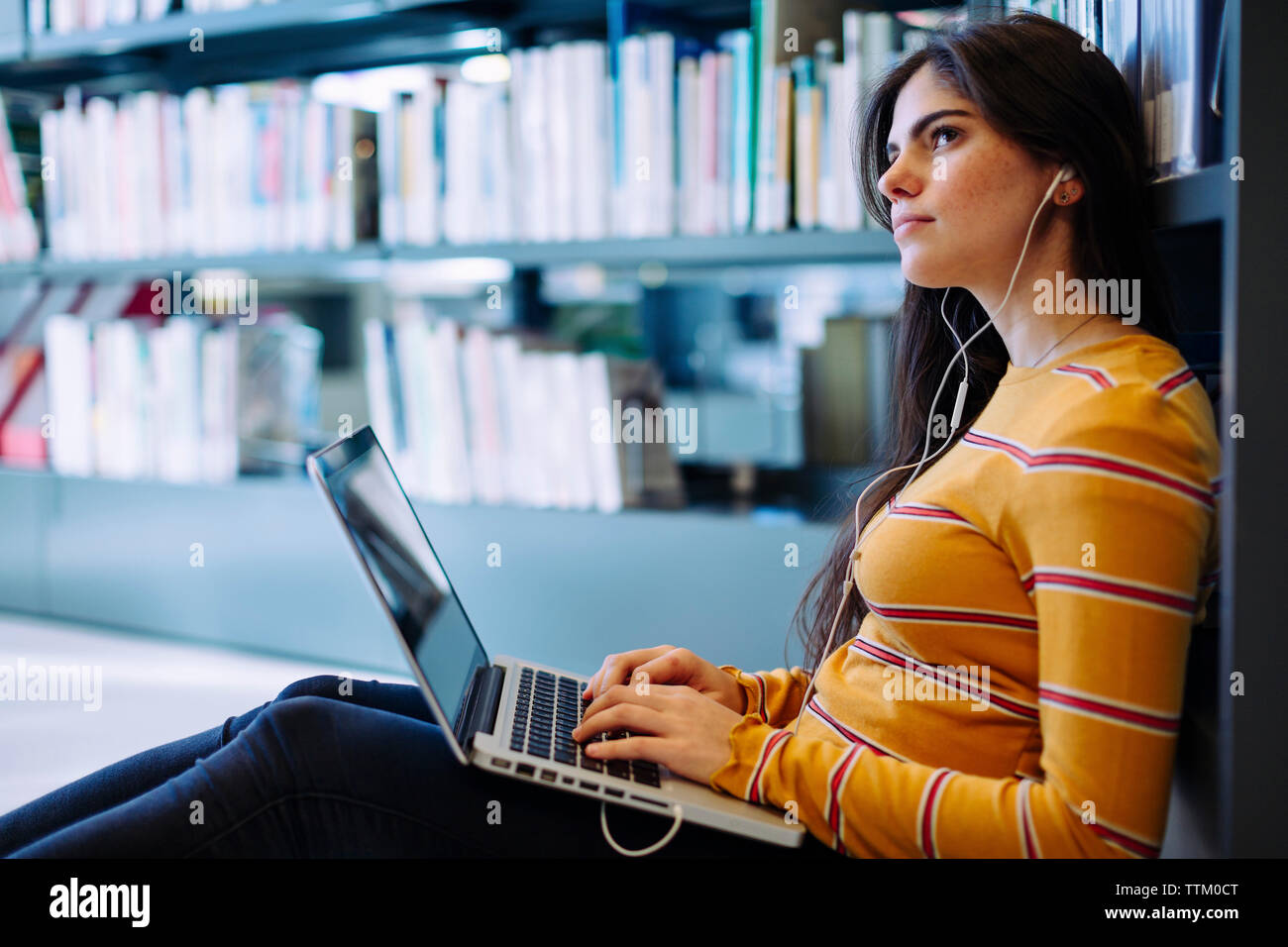 Nachdenkliche Frau mit Laptop sitzen durch Bücherregale in Bibliothek Stockfoto