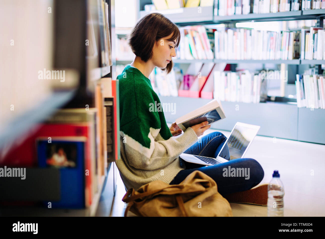 Seitenansicht der Frau mit Laptop lesen Buch beim Sitzen auf dem Boden in der Bibliothek Stockfoto