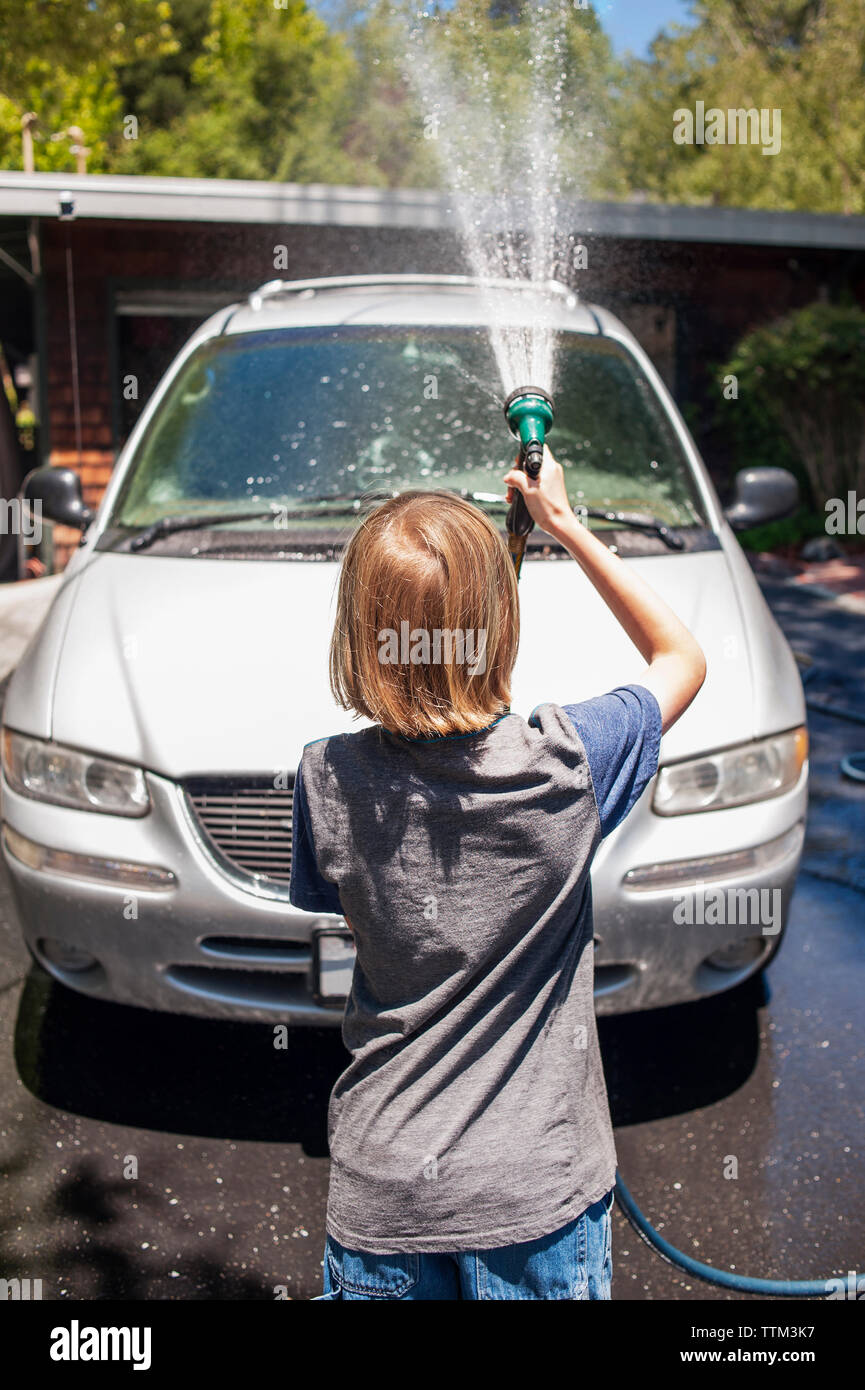 Ansicht der Rückseite des Jungen waschen Auto mit Gartenschlauch in Einfahrt Stockfoto