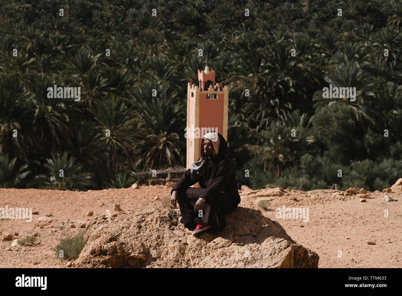 Mann mit geschlossenen Augen tragen traditionelle Kleidung beim Sitzen auf Rock gegen Palmen in der Sahara. Stockfoto