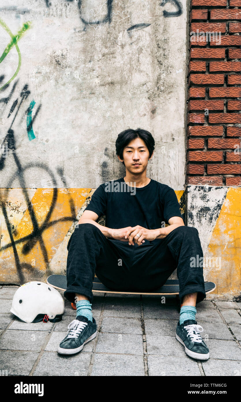 Portrait von ernsten Mann sitzt auf Skateboard gegen die Wand in der Stadt Stockfoto