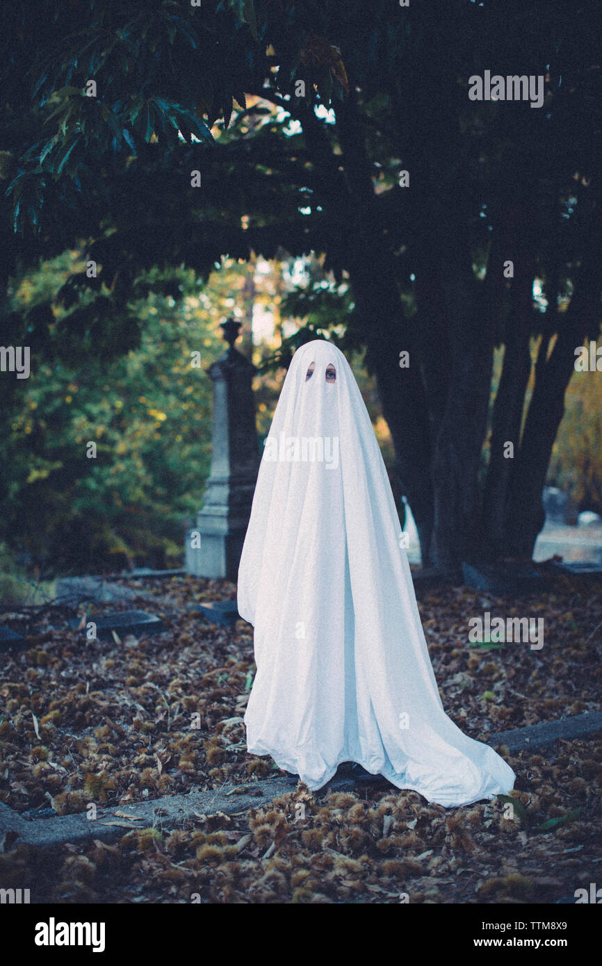 In voller Länge Porträt des jungen tragen ghost Kostüm beim Stehen auf Feld am Friedhof während der Halloween Stockfoto