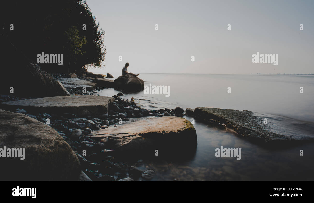 Seitenansicht der Junge sitzt auf Felsen am Ufer gegen Sky Stockfoto