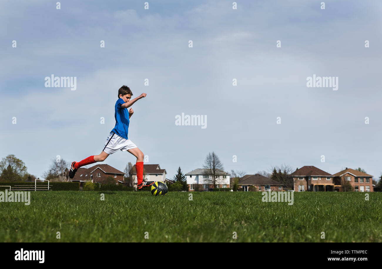 Seitenansicht des Jungen kicken Fußball beim Spielen auf Wiese gegen Sky Stockfoto