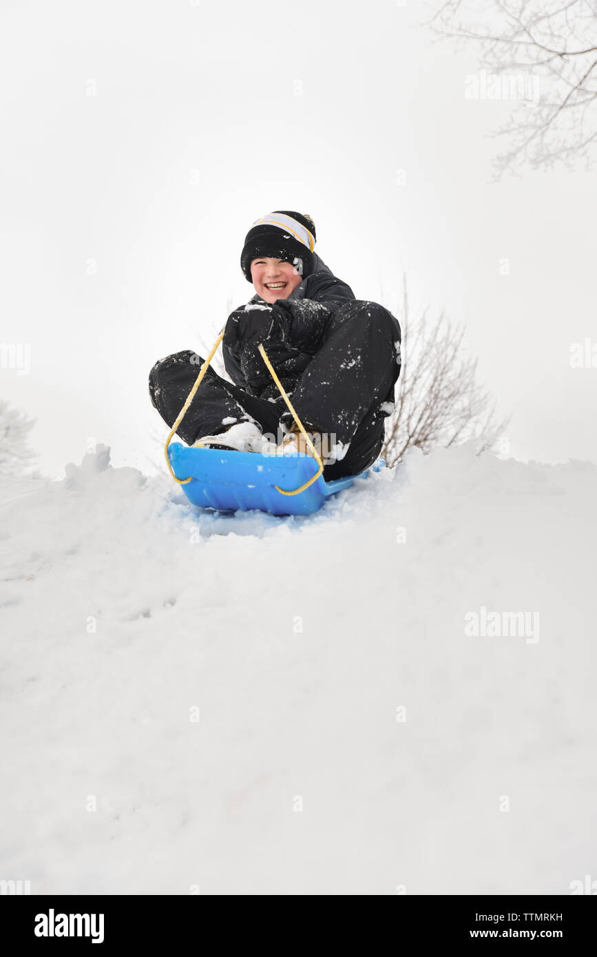 Lächelnde Junge sitzt auf einer Rodelbahn auf verschneiten Hügel an einem Wintertag. Stockfoto