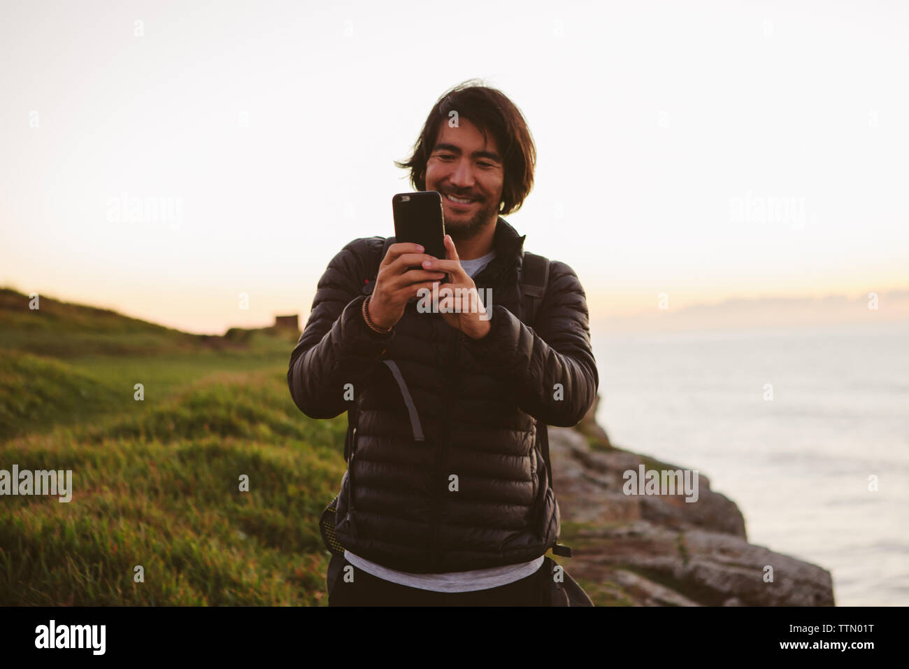 Gerne männliche Wanderer Fotografieren durch Smart Phone beim Stehen auf einem Hügel mit Meerblick gegen den klaren Himmel Stockfoto