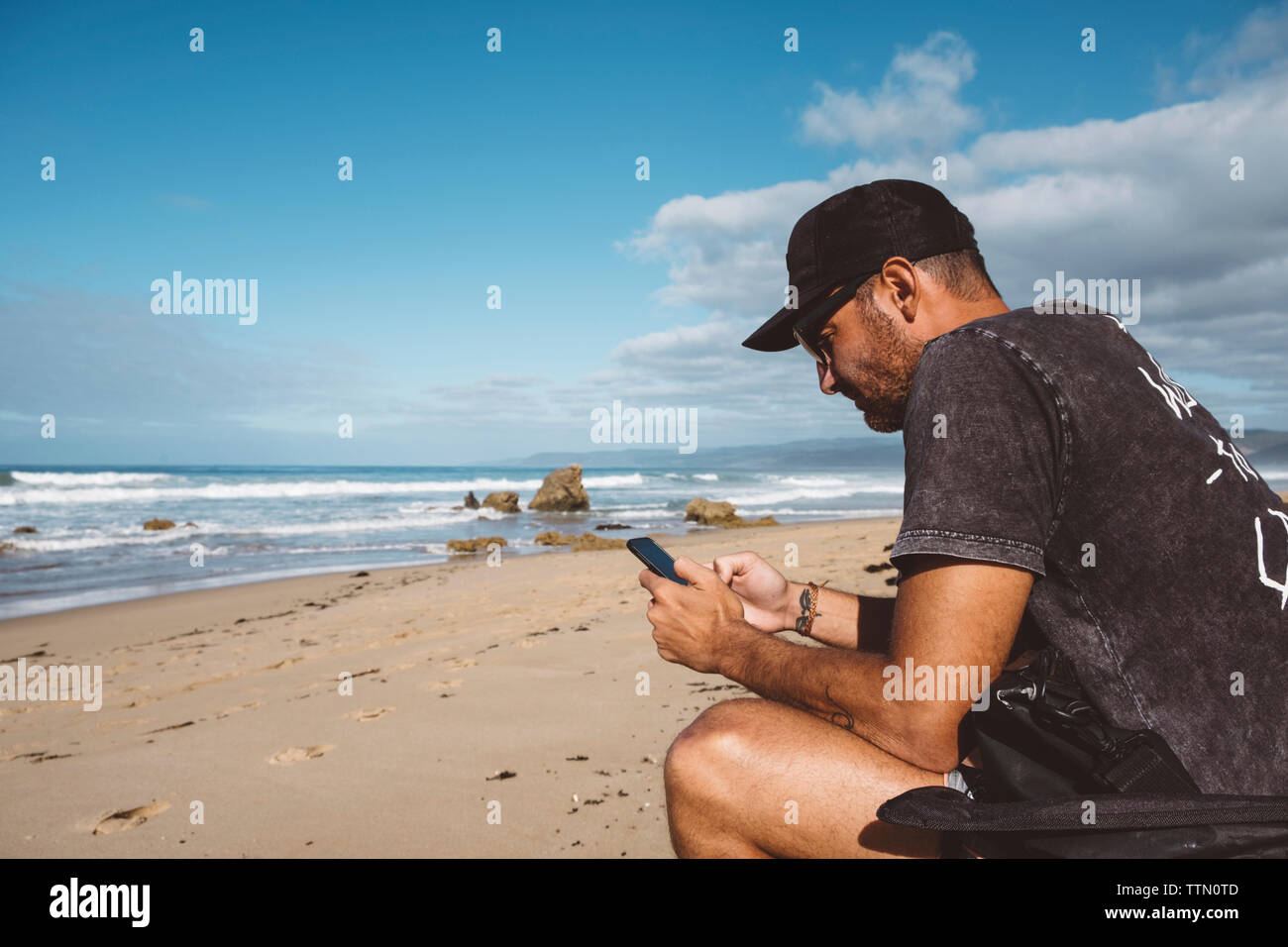 Seitenansicht der Mann mit Handy, während am Strand gegen den blauen Himmel sitzen während der sonnigen Tag Stockfoto