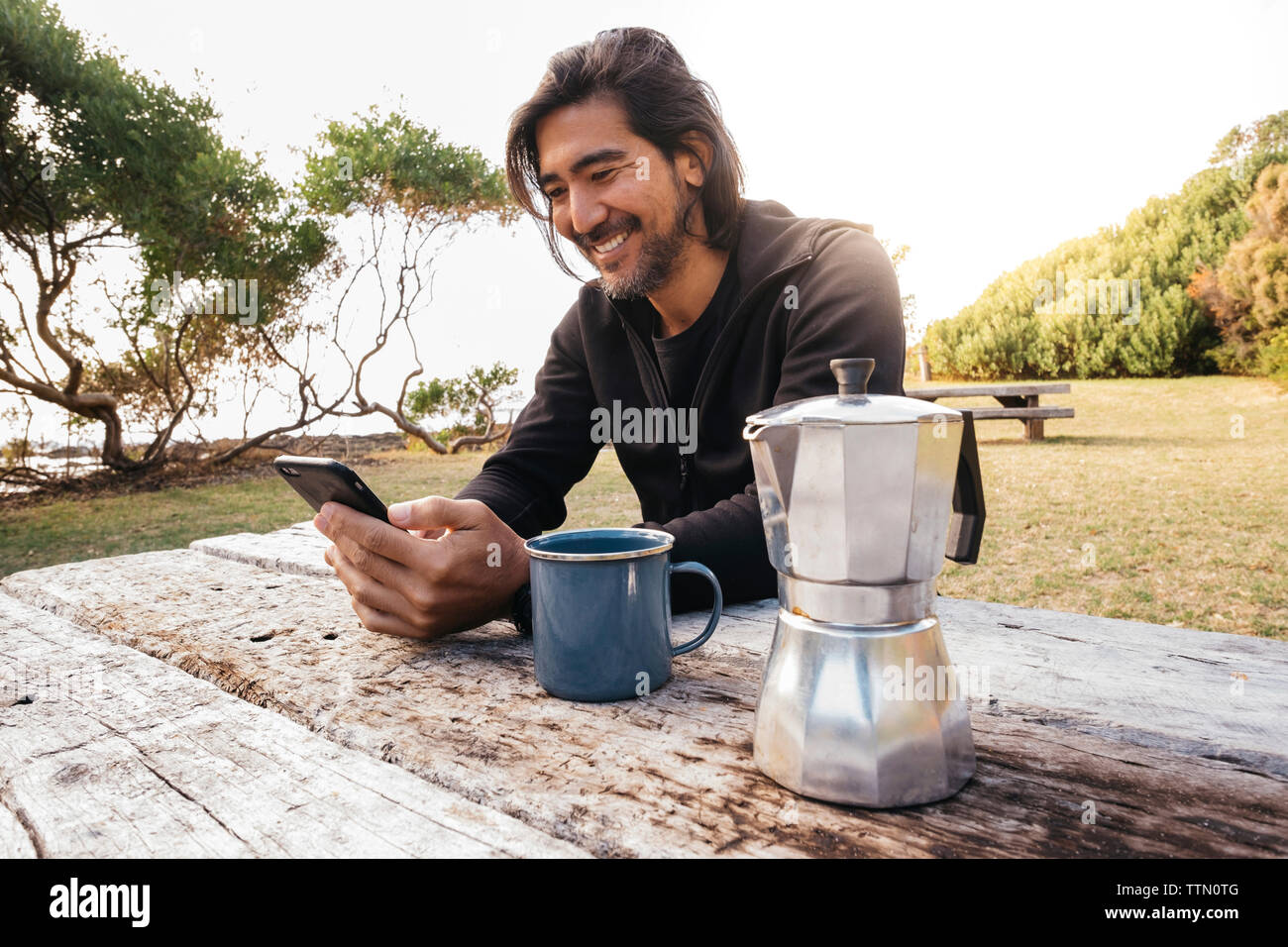Glückliche Menschen mit Kaffee auf Holztisch mit Handy während gegen Himmel sitzen Stockfoto