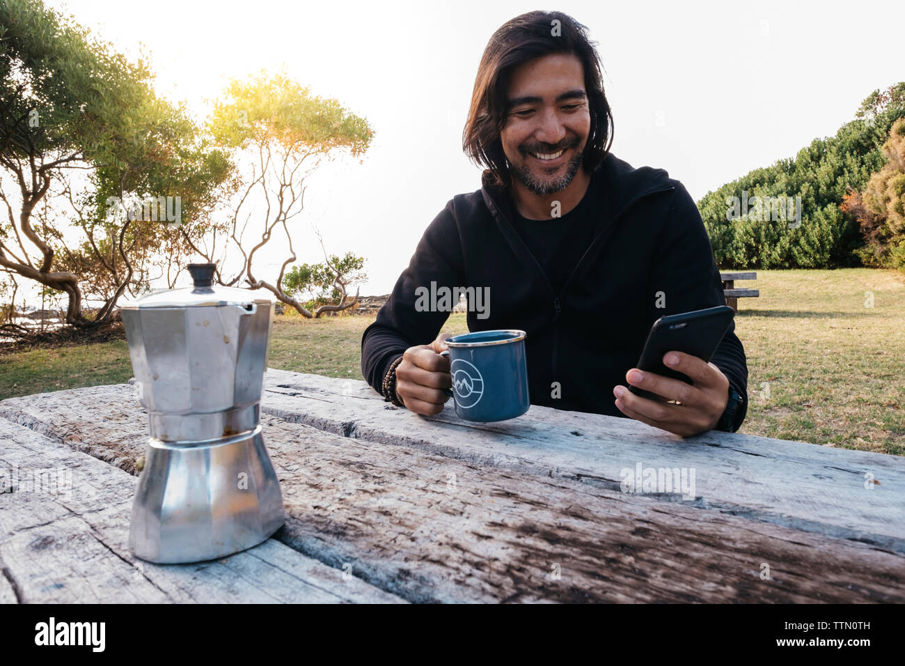 Glücklicher Mann mit Handy Kaffee auf hölzernen Tisch sitzend gegen Sky Stockfoto