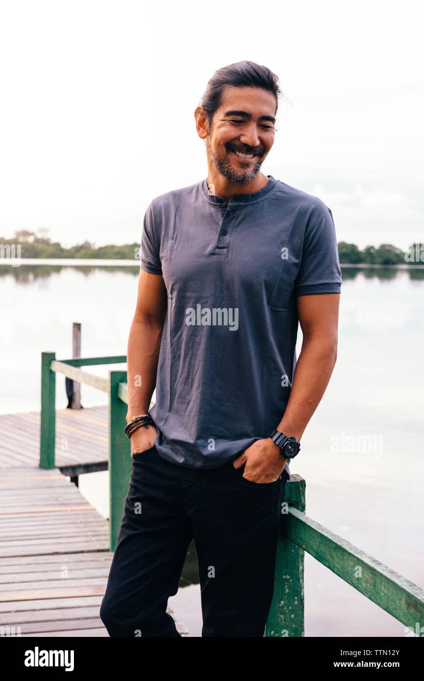 Lächelnd reifer Mann mit Händen in den Taschen auf Pier gegen See Stockfoto