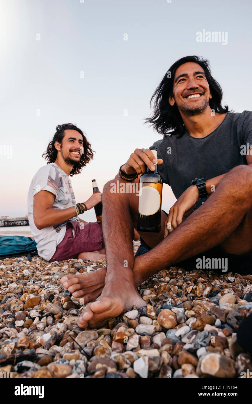 Lächelnd Freunde mit Alkohol Flaschen sitzen auf Kieselsteinen am Strand gegen Sky Stockfoto
