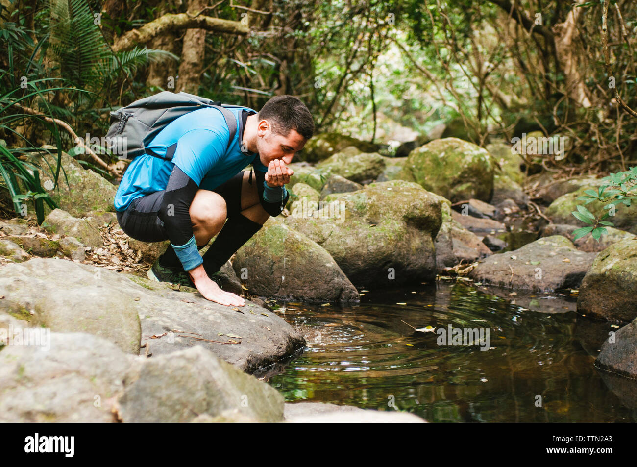 Männliche Wanderer Trinkwasser aus See während kauert auf Felsen im Wald Stockfoto