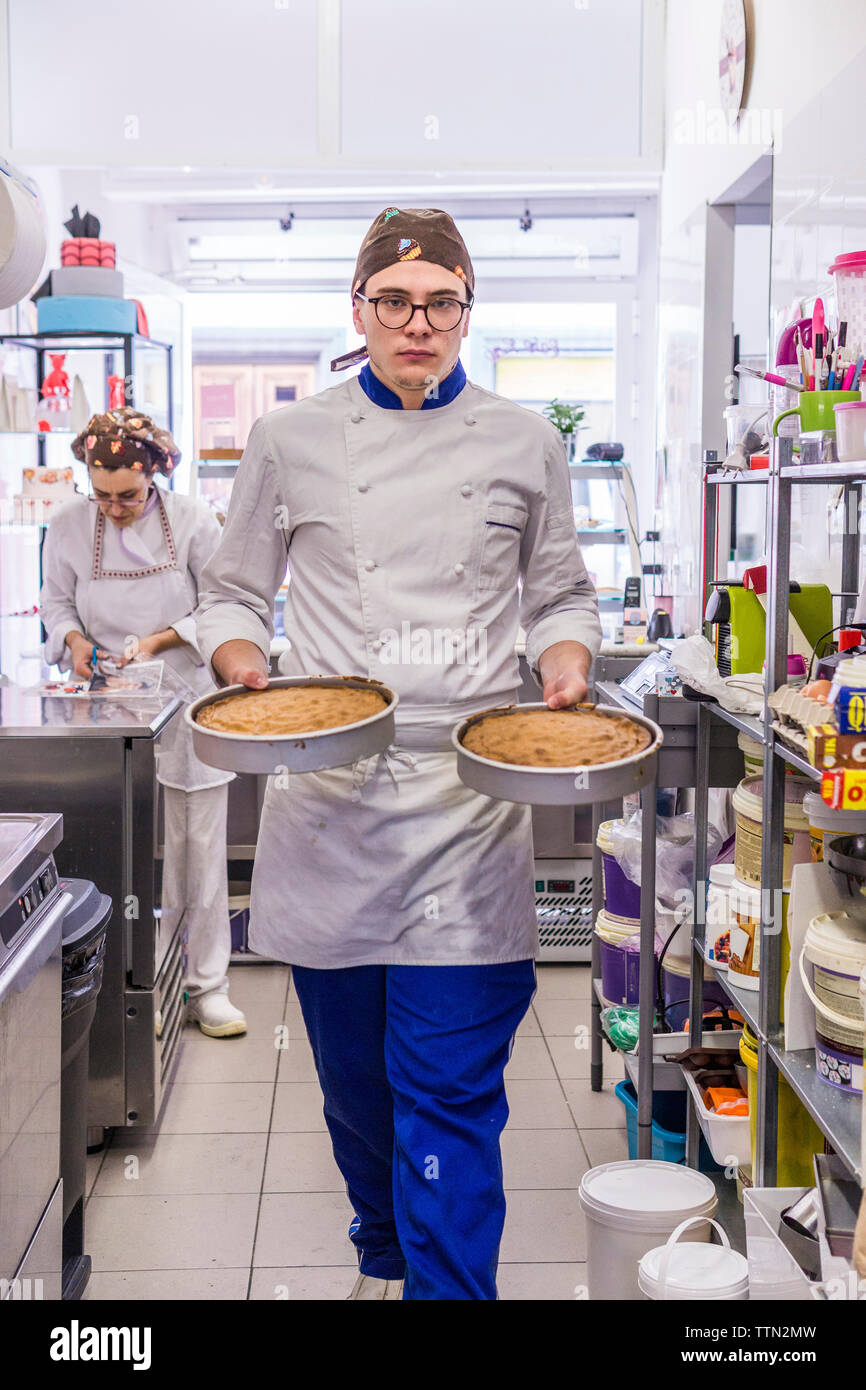 Portrait der männlichen Bäcker Kuchen halten, während die weiblichen Mitarbeiter arbeiten im Hintergrund bei Labor Stockfoto