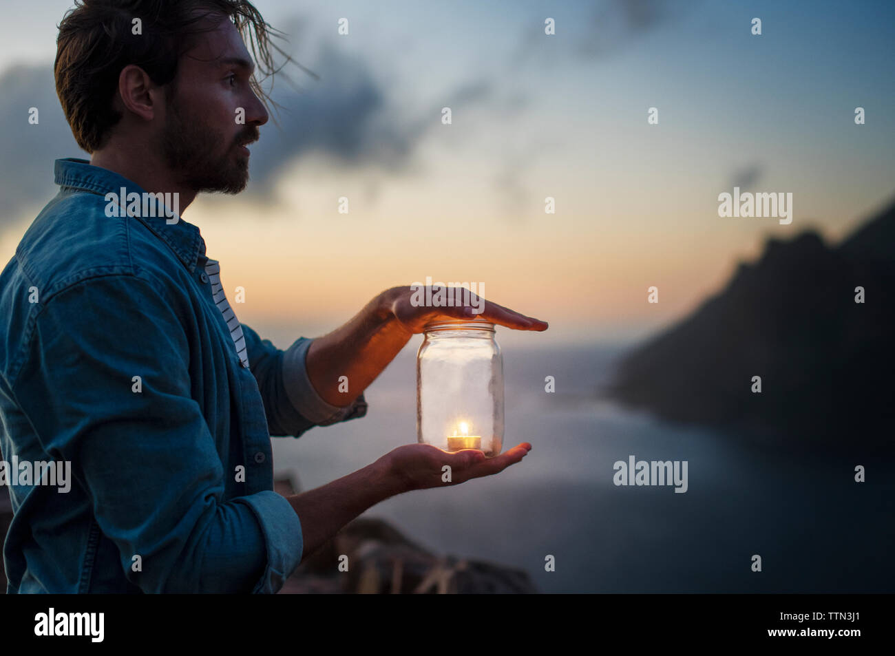 Seitenansicht der Mann hält Teelicht Kerze im Glas durch das Meer bei Sonnenuntergang Stockfoto
