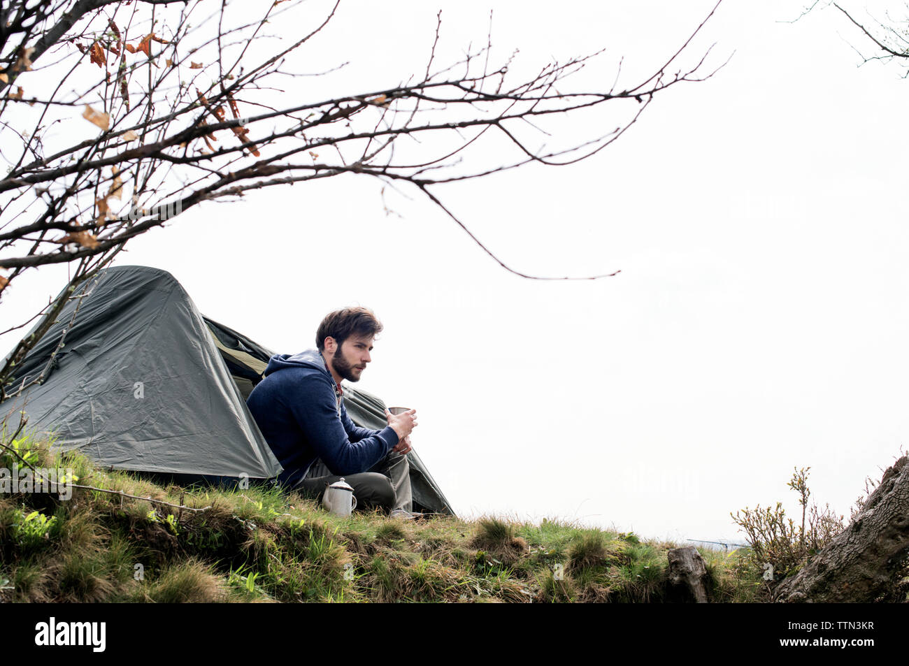 Nachdenklicher Mann sitzt außerhalb Zelt auf Hügel gegen klaren Himmel Stockfoto