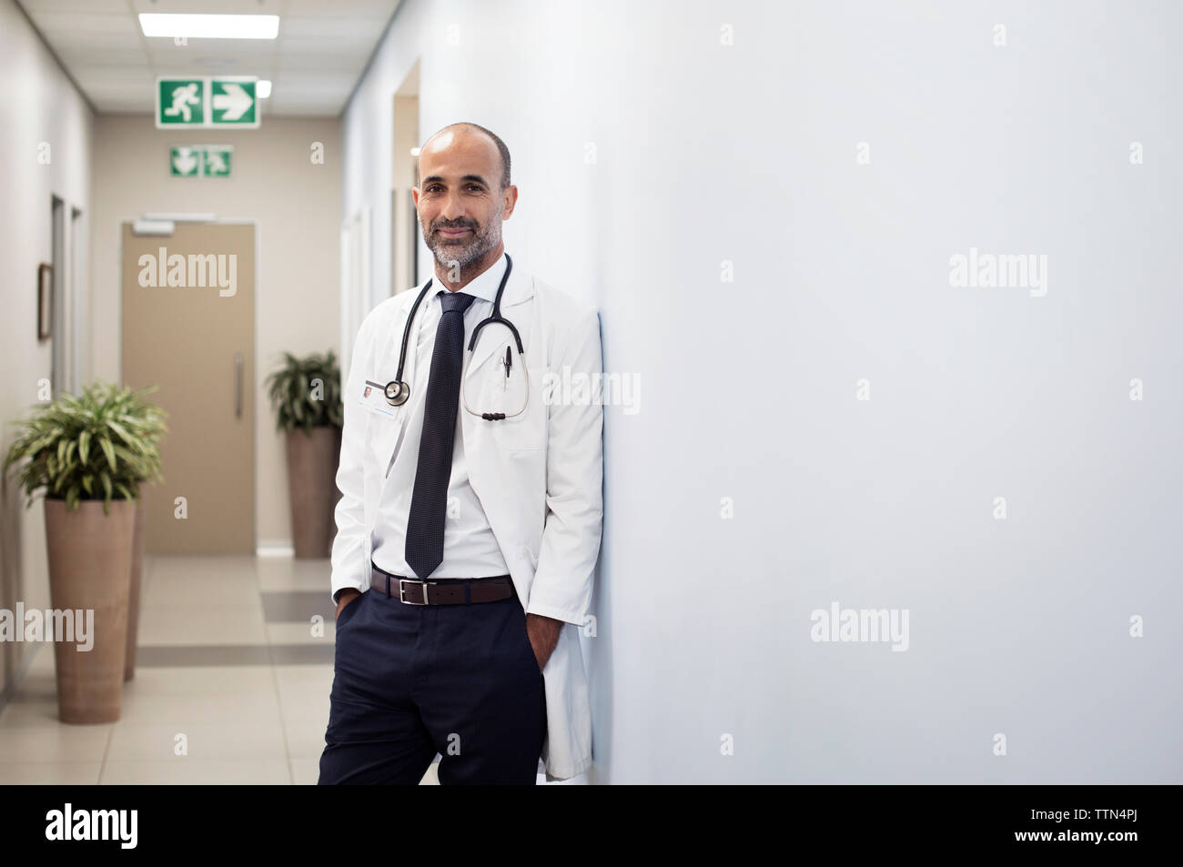 Portrait von lächelnden Arzt mit Händen in den Taschen an der Wand am Krankenhaus schiefen Stockfoto