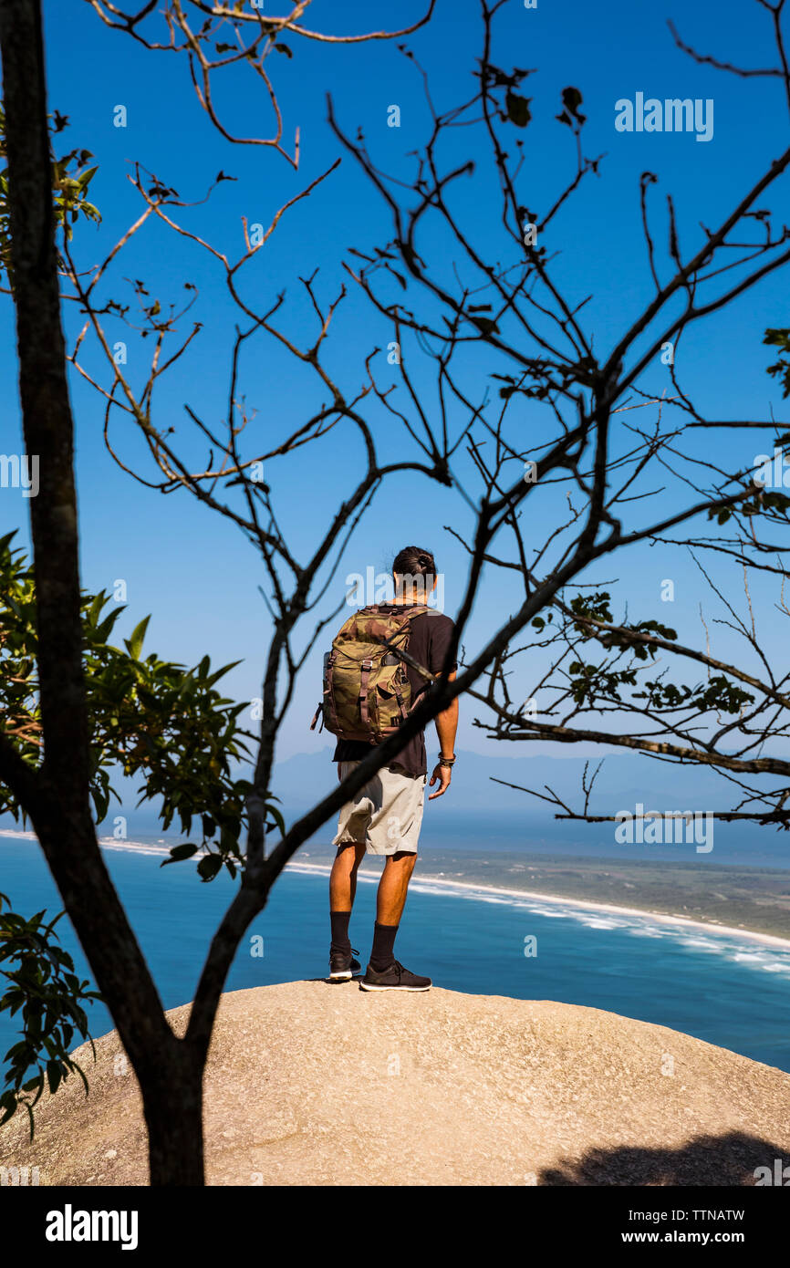 Volle Länge des Backpacker suchen auf See beim Stehen auf Klippe gegen den blauen Himmel Stockfoto