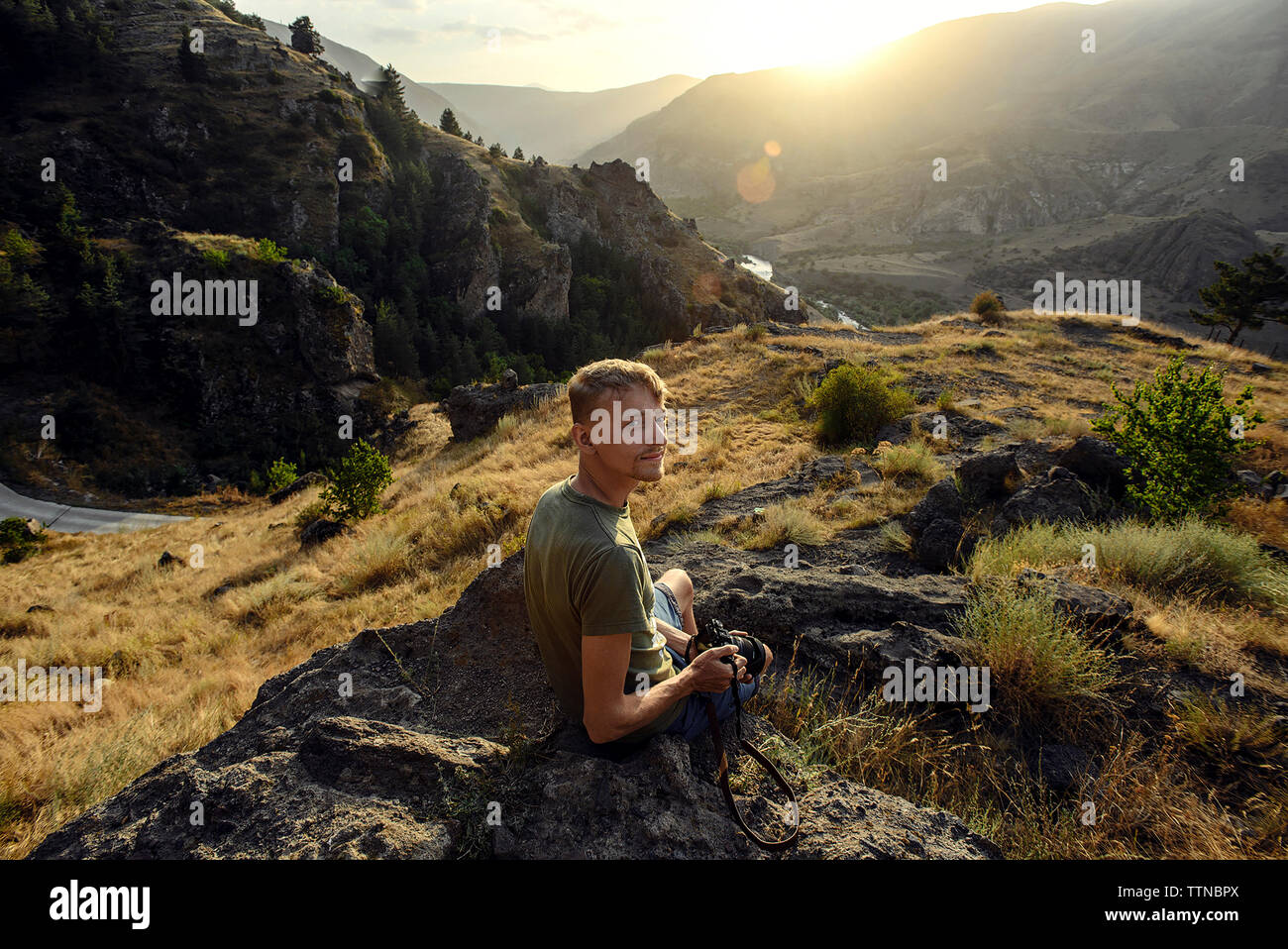 Porträt der Mann mit der Kamera sitzen auf Rock gegen die Berge bei Sonnenuntergang Stockfoto
