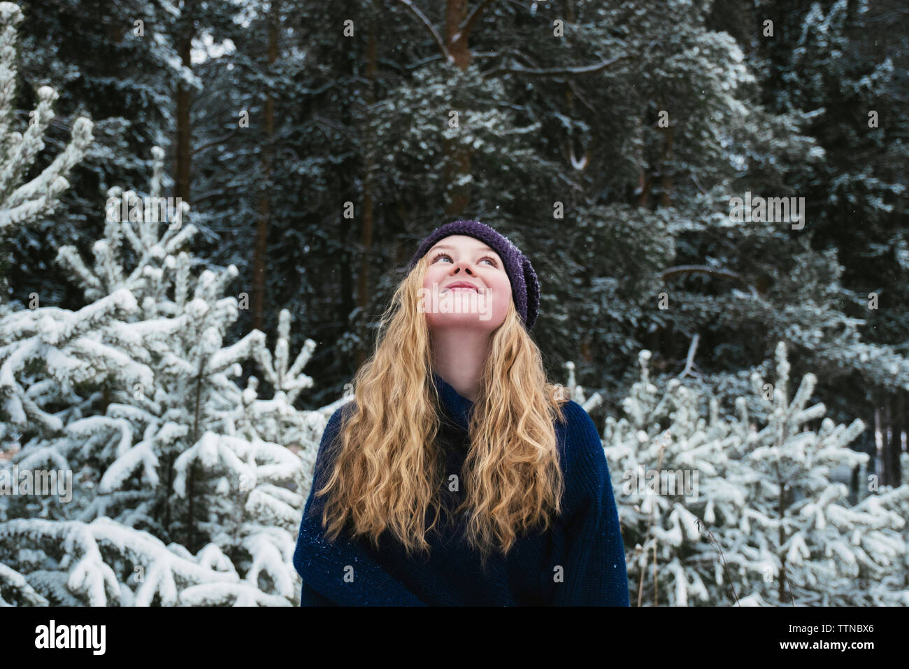 Glückliche Frau mit blonden Haaren, die beim Stehen gegen Bäume im Wald im Winter Stockfoto