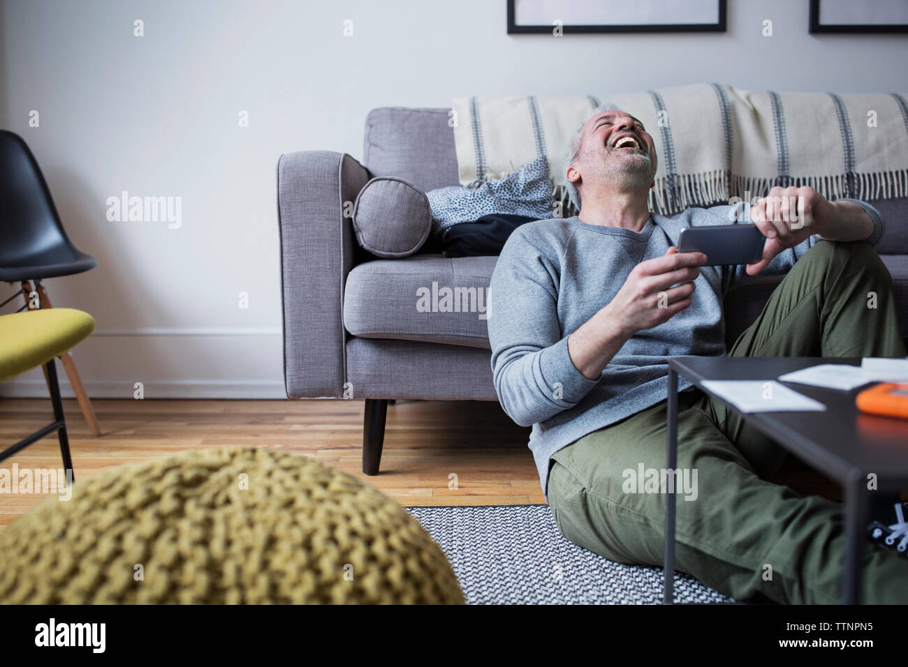 Fröhliche reifer Mann Holding smart phone beim Sitzen auf dem Fußboden im Wohnzimmer. Stockfoto