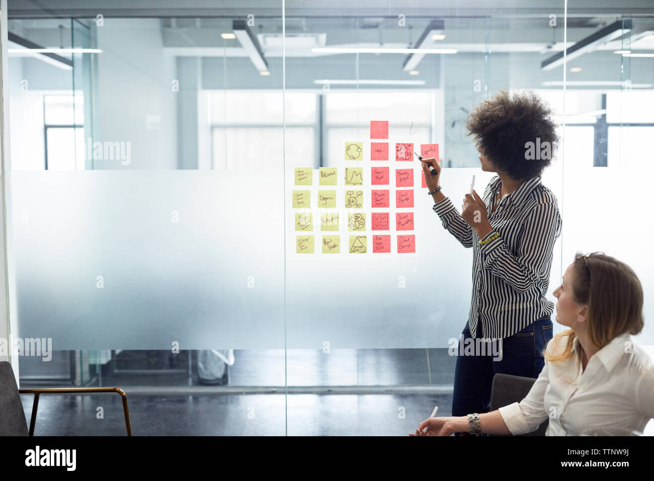 Geschäftsfrau mit Klebenotizen an Glaswand vor einer Kollegin im Büro Stockfoto