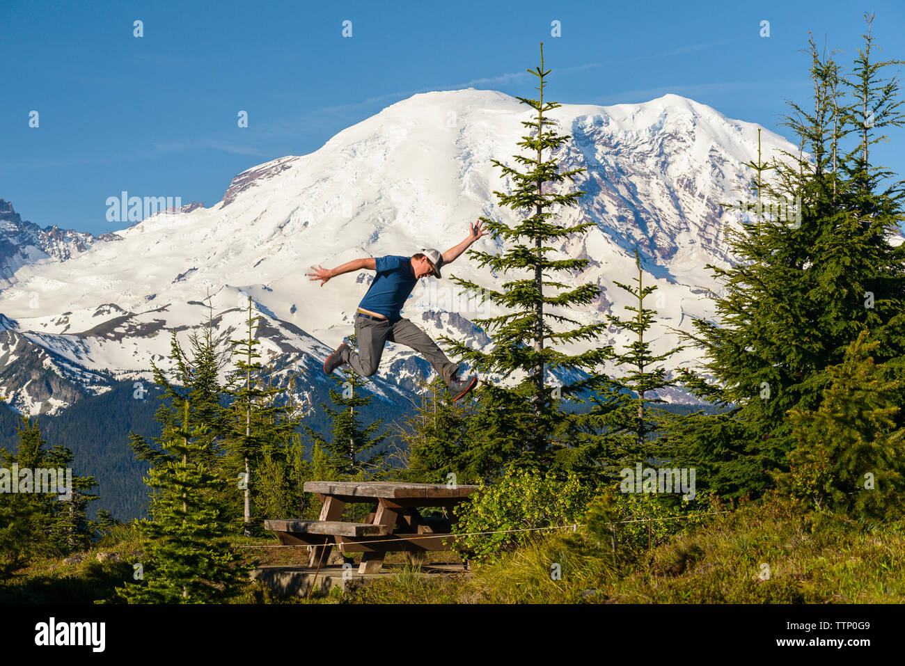 Mann springen über Picknicktisch im Feld gegen schneebedeckte Berge begeistert Stockfoto