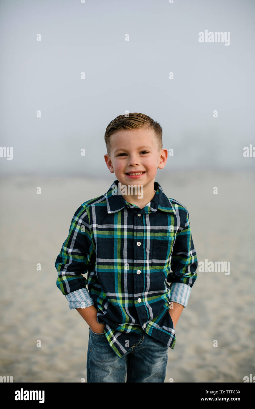 Portrait von süßen Jungen mit Händen in den Taschen am Strand gegen Himmel während nebligen Wetter Stockfoto