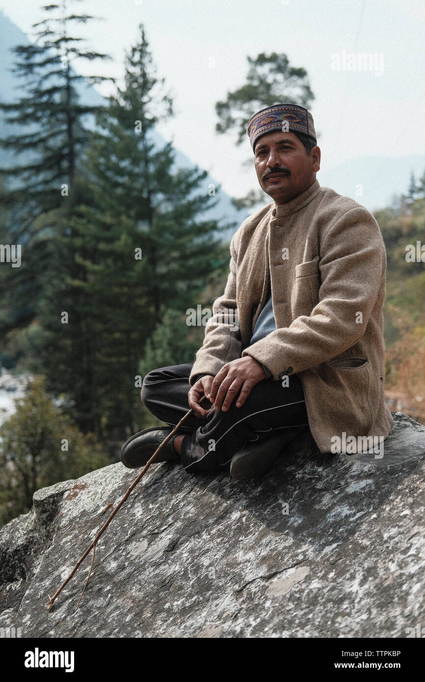 Die volle Länge der reifen Mann, der warme Kleidung holding Stick beim Sitzen auf Rock im Wald Stockfoto
