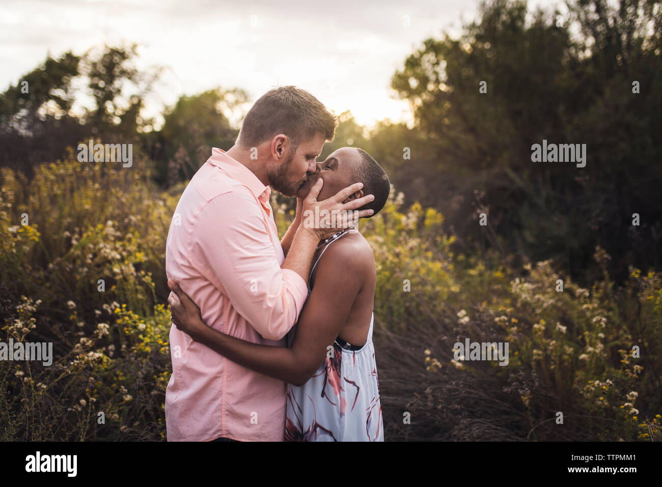 Seitenansicht des Romantisches Paar küssen beim stehen inmitten von Pflanzen gegen Himmel im Park bei Sonnenuntergang Stockfoto