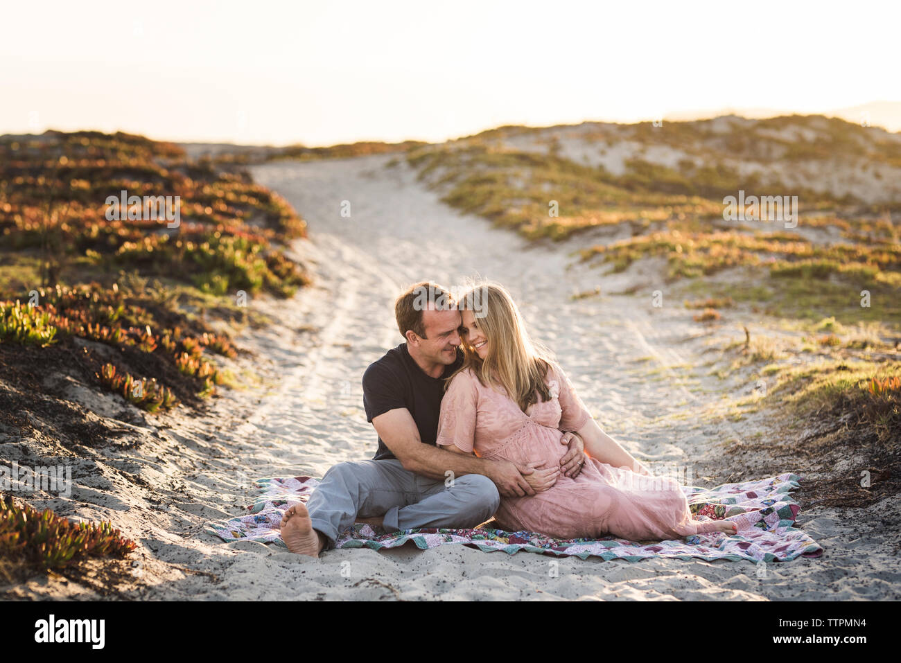 Romantisches Paar sitzt auf Decke am Strand gegen den klaren Himmel bei Sonnenuntergang Stockfoto