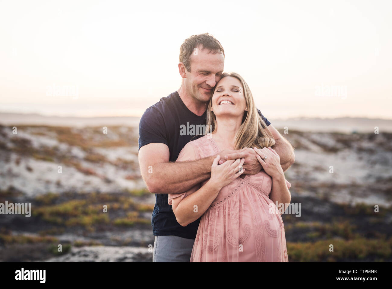 Romantisches Paar umarmt, während am Strand gegen den klaren Himmel stehen bei Sonnenuntergang Stockfoto