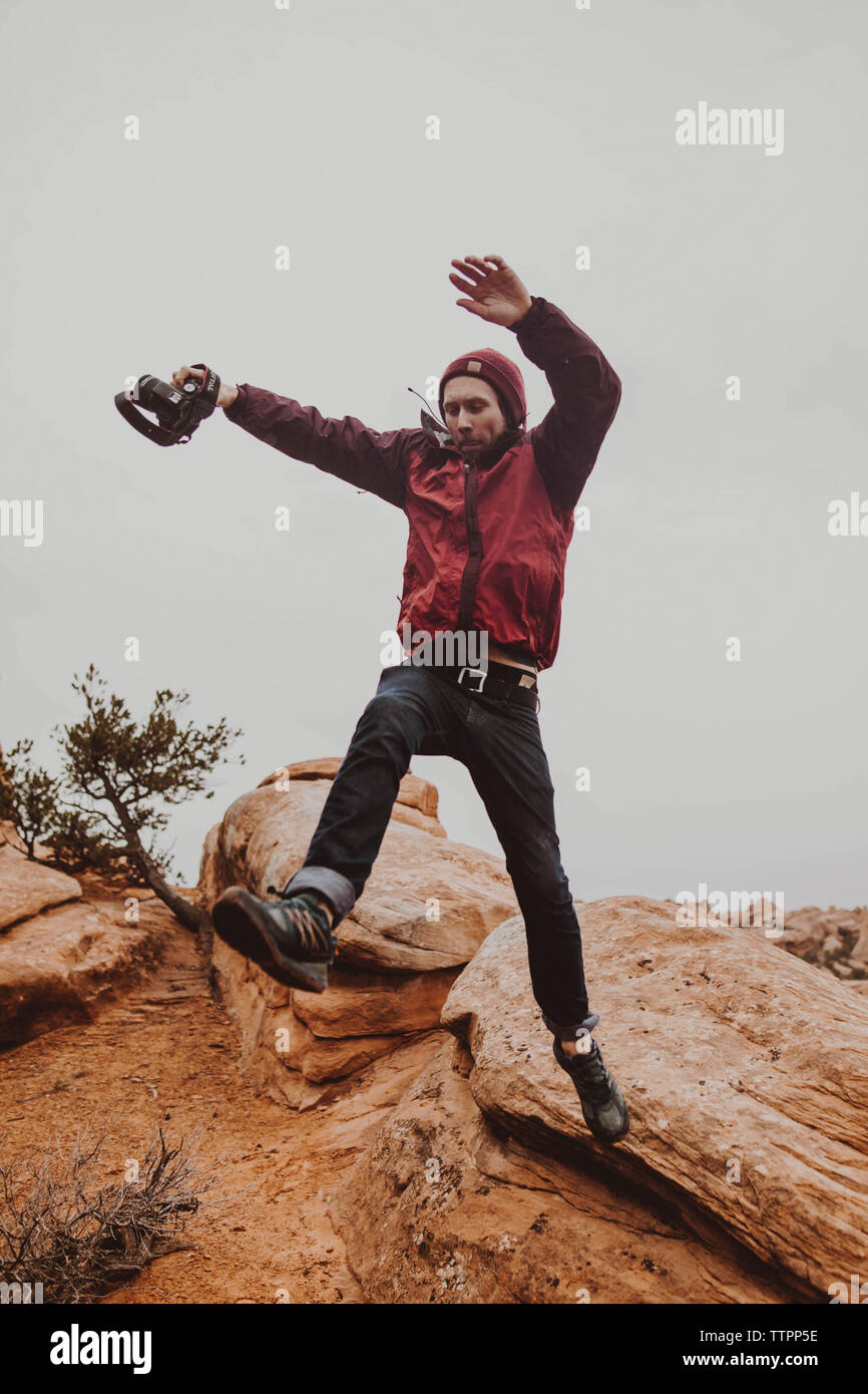 Low Angle View der Mann hält Kamera beim Springen auf den Felsen gegen Sky Stockfoto