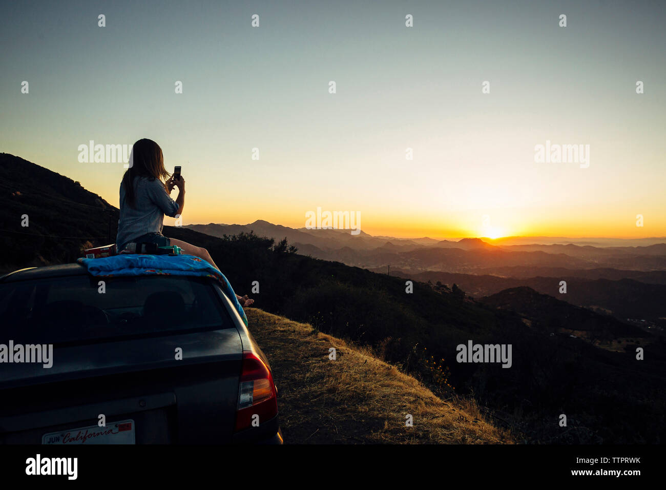 Seitenansicht der Frau fotografieren Berge mit Handy beim Sitzen auf dem Dach gegen den klaren Himmel bei Sonnenuntergang Stockfoto
