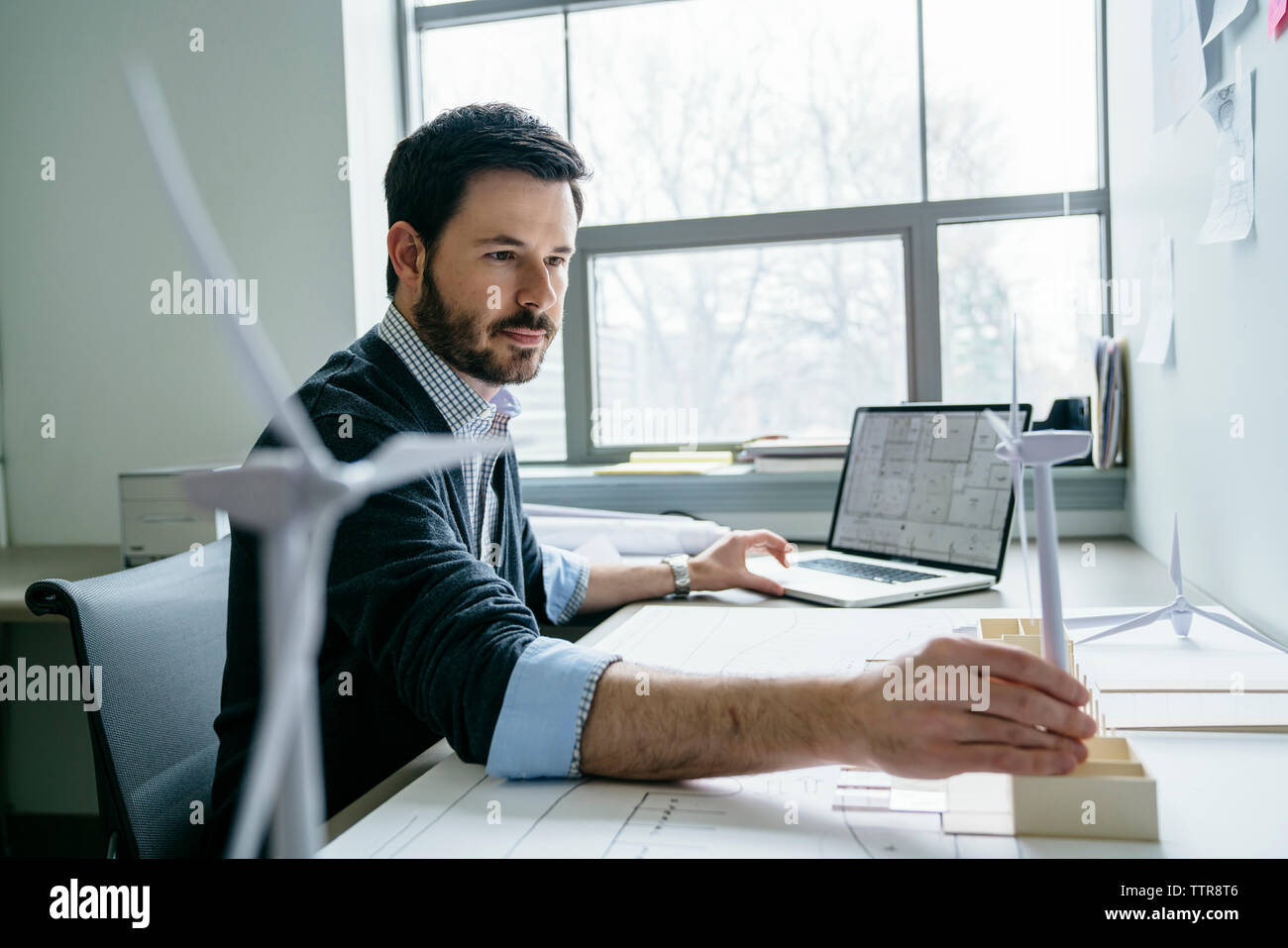 Geschäftsmann, der ein Notebook benutzt, während er das Modell einer Windkraftanlage auf dem Schreibtisch arrangiert Stockfoto