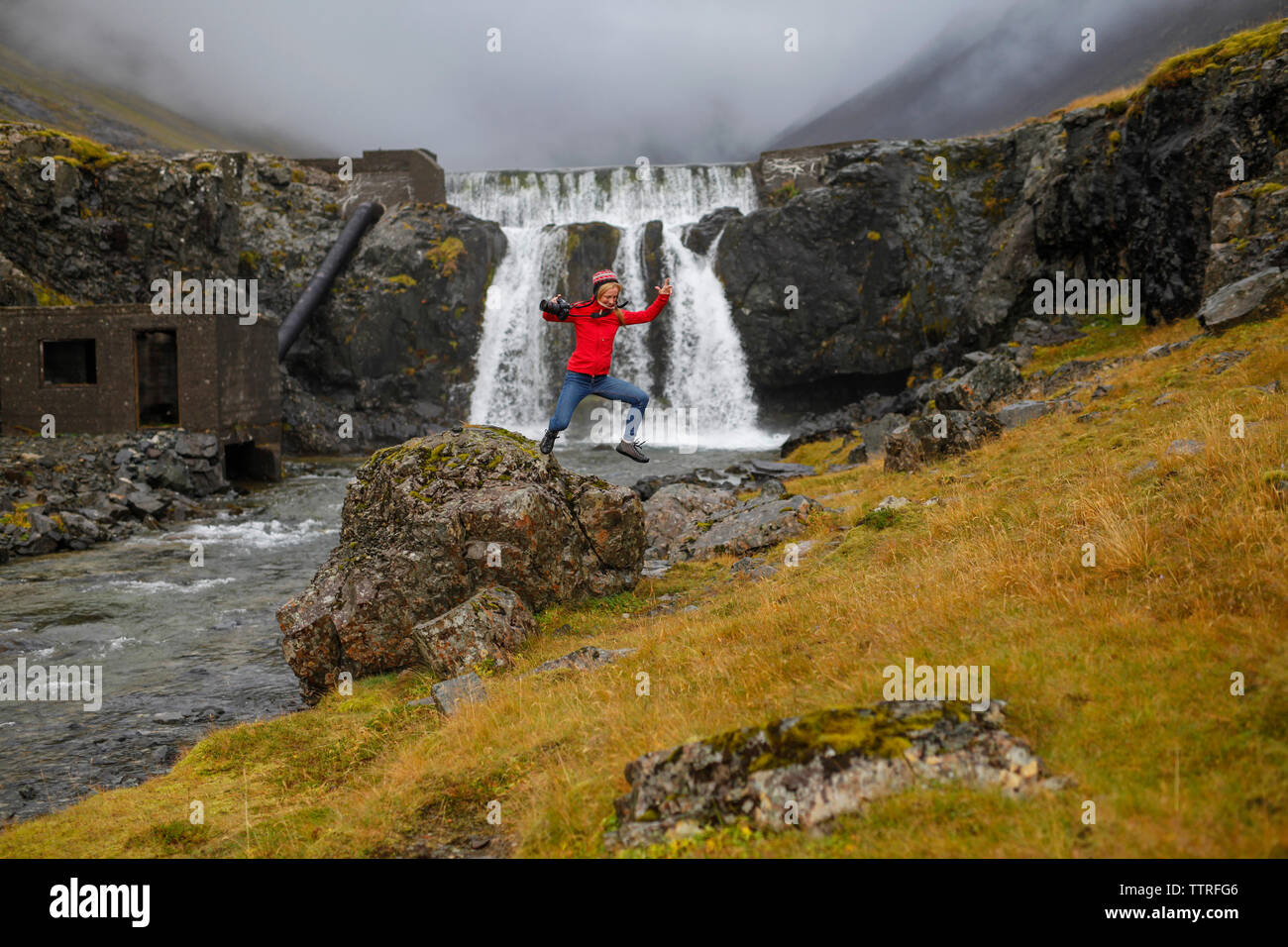 Weibliche Wanderer das Springen von Rock gegen Wasserfall aufgeregt Stockfoto