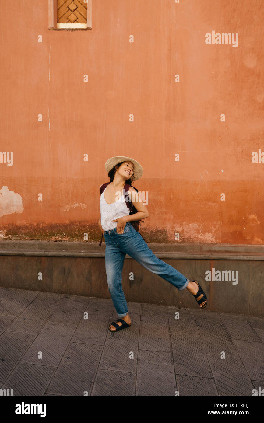 Volle Länge des aufgeregt weibliche Touristen mit Rucksack und die Hände in den Taschen tanzen gegen die Wand in der Stadt Stockfoto