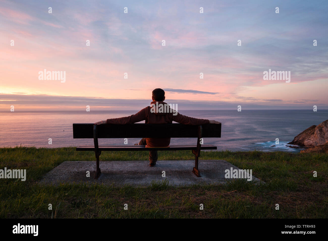 Volle Länge des Menschen mit Blick auf das Meer beim Sitzen auf der Bank gegen bewölkter Himmel bei Sonnenuntergang Stockfoto