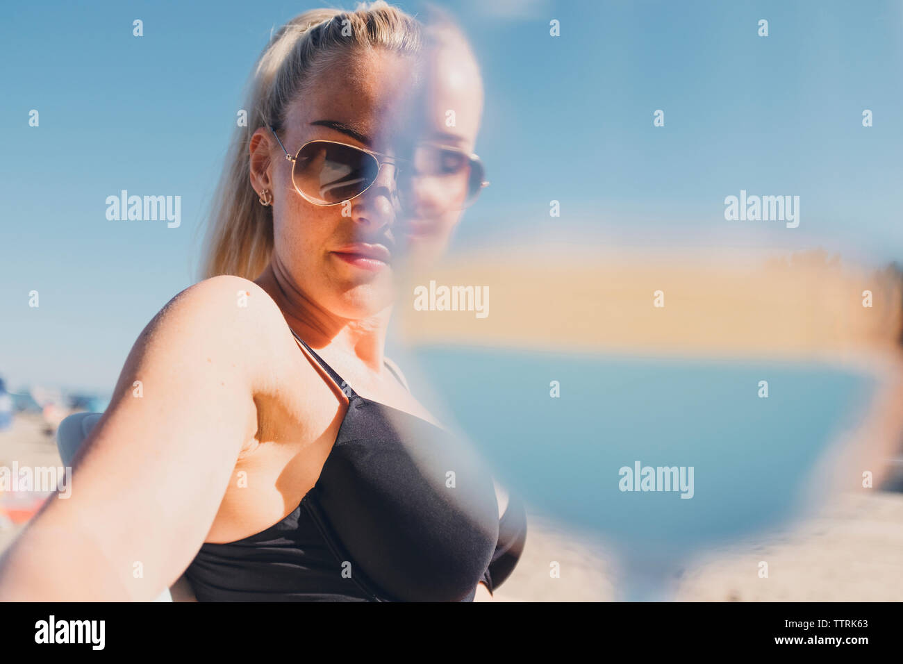 Portrait von Frau mit Sonnenbrille und Bikini am Strand mit Glas im Vordergrund Stockfoto
