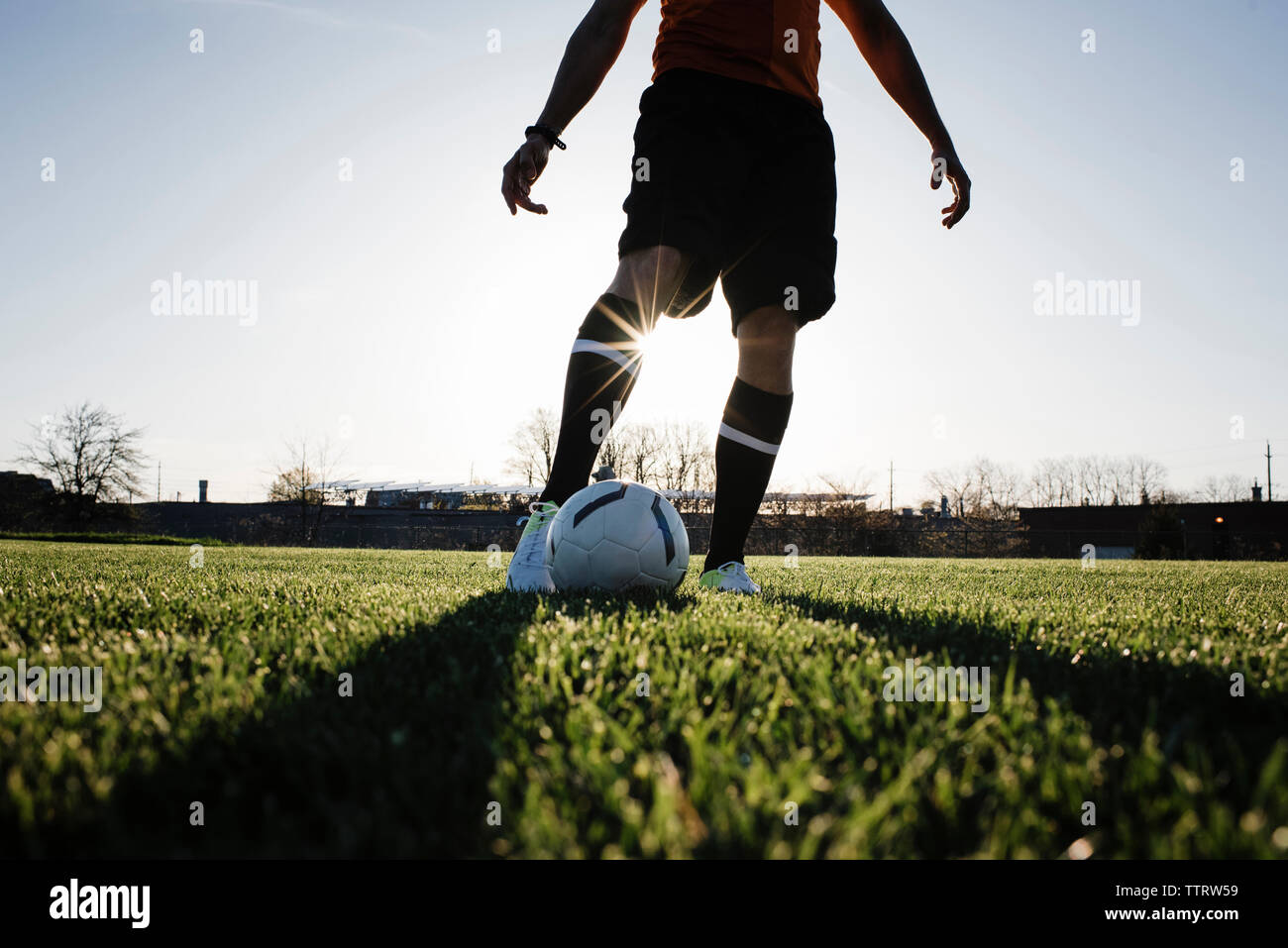 Untere Partie der Mann spielt Fußball auf der Wiese gegen den klaren Himmel bei Sonnenuntergang Stockfoto