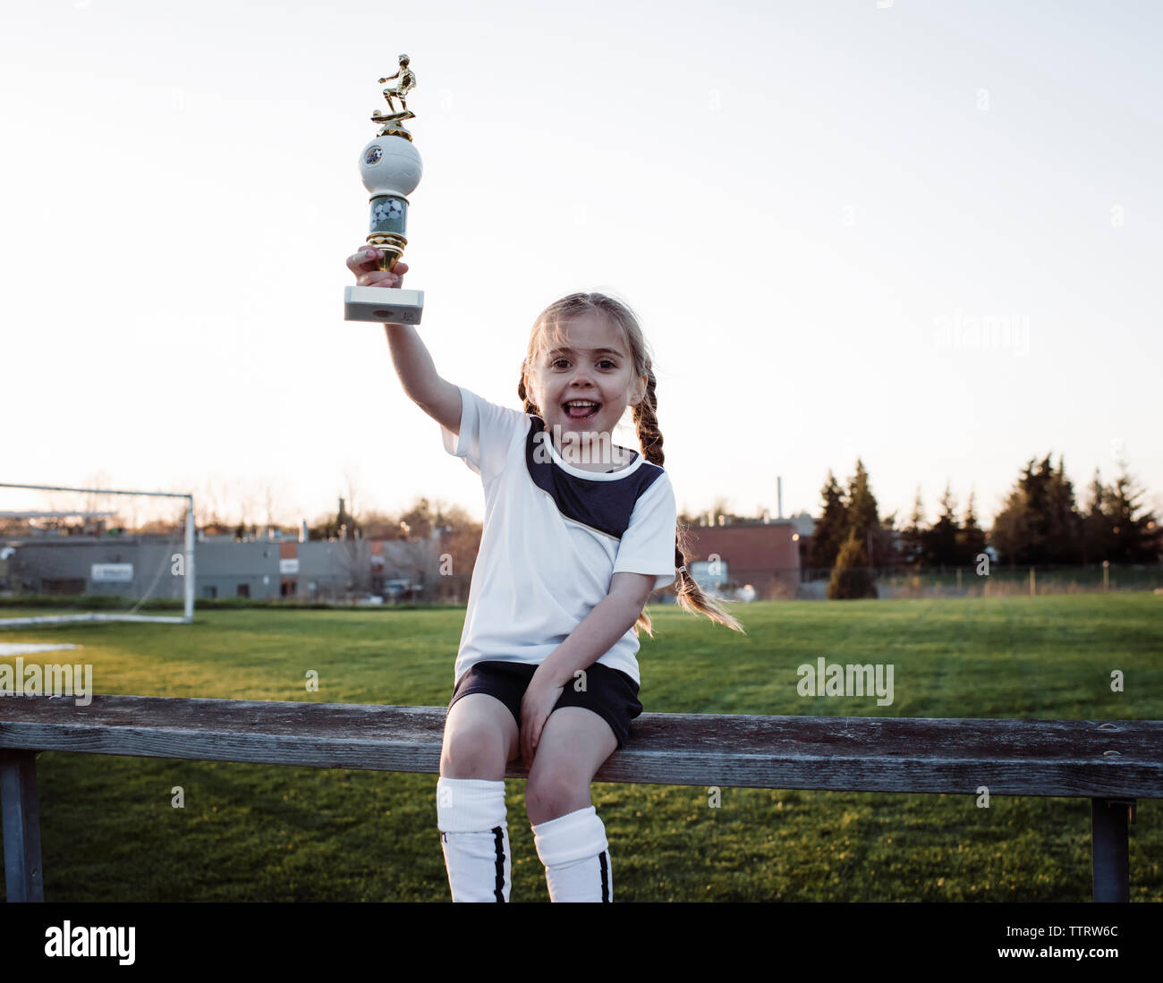 Portrait von Happy girl Holding Trophäe beim Sitzen auf der Bank gegen den klaren Himmel im Park Stockfoto